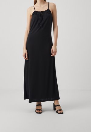 Femme portant une robe noire sans manches, longue jusqu'au sol, avec des fines bretelles et un détail noué à l'avant, associée à des sandales noires à talons hauts.
