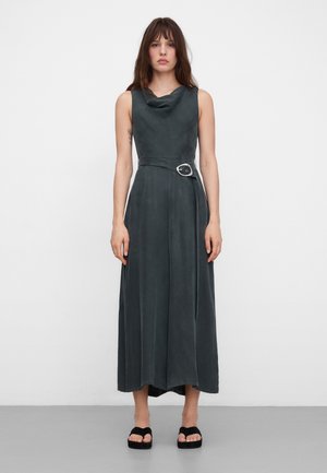 Kobieta stojąca, ubrana w ciemnozieloną sukienkę midi bez rękawów z dekoltem typu komin i paskiem z owalną klamrą, czarne sandały na platformie, na jednolitym białym tle.