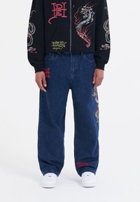 Baggy blå denimjeans med röd och flerfärgad brodyr, med detaljer på framsidan och sidan. Bärs med en svart zip-up hoodie.