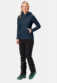 Granatowa pikowana kurtka z przednim zamkiem błyskawicznym, dwiema bocznymi kieszeniami; w połączeniu z czarnymi spodniami i zielonymi butami narciarskimi; zaprojektowana do aktywności na świeżym powietrzu.