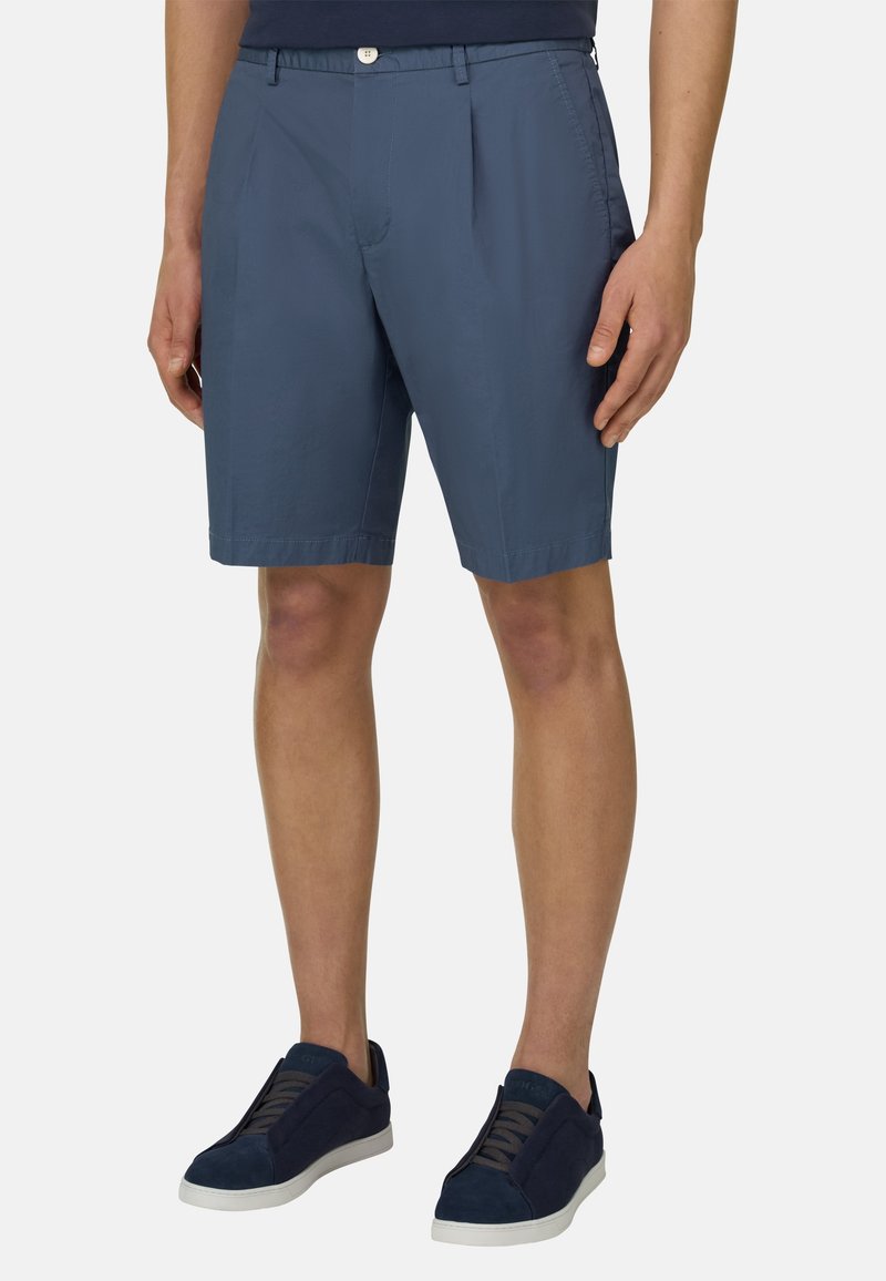 Homme portant un short bleu mi-long avec des plis à l'avant et des baskets bleu marine avec des semelles blanches, debout devant un fond uni.