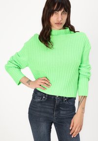 Pulover tricotat de culoare verde lime strălucitor, cu guler înalt și textură ribbed, asortat cu blugi negri.