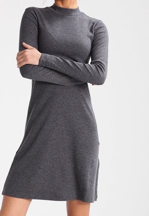 Mujer con un vestido de punto gris oscuro, de manga larga y largo hasta la rodilla, con cuello alto, parada con los brazos cruzados frente a un fondo blanco.