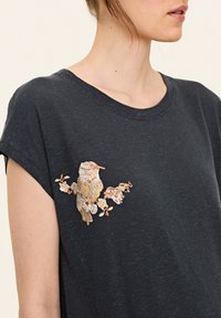 Schwarzes T-Shirt mit kurzen Ärmeln, das ein gold- und cremefarbenes besticktes Vogelmotiv mit floralen Akzenten auf der linken Seite zeigt. Weicher Stoff, lässige Passform.