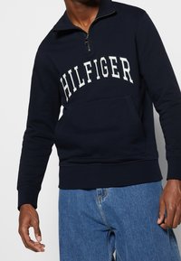 Homme portant un sweat-shirt marine à demi-fermeture éclair avec le texte "HILFIGER" sur le devant et un jean bleu, debout devant un fond clair uni.