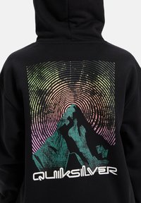 Černá mikina s kapucí s retro grafikou hor ve teal barvě a koncentrovanými vzory kruhů v zelené, růžové a fialové. Název značky v bílé barvě.