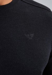 Tmavě námořnický pletený svetr s kulatým výstřihem a akcenty na ramenou, se subtilním vyšitým logem na hrudi. Jemná textura.