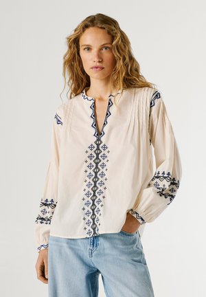 Woman with wavy blonde hair wearing a cream blouse with blue geometric embroidery and light blue jeans, posing with one hand in pocket.