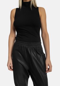 Sort ribbet turtle neck top med ærmeløs design, parret med sorte, blanke bukser med elastik i taljen og sidelommer.