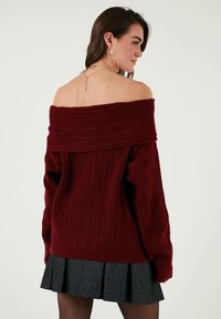 Pulover burgundy cu dungi, fără umeri, cu un guler lat și mâneci lungi, purtat peste o fustă plisată neagră. Model tricotat texturat vizibil.
