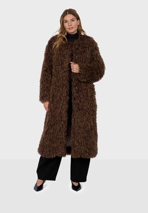 Femme portant un long manteau en fourrure texturée marron foncé sur un pantalon noir et des chaussures pointues noires, debout devant un fond clair uni.