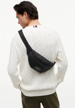 Uomo che indossa un maglione bianco a trecce e pantaloni verdi con una borsa a tracolla nera portata sulla schiena.