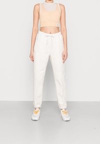 Osoba w beżowym topie na krótko, białych dresach z elastycznym pasem i białych trampkach z żółtymi sznurówkami stoi na tle jednolitego tła.