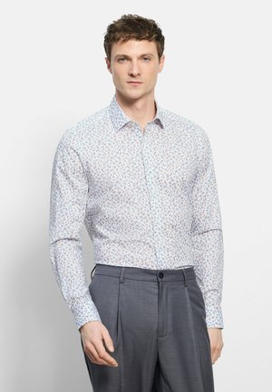 Jonge man draagt een wit overhemd met lange mouwen met kleine kleurrijke bloemmotieven, ingestopt in een grijze broek met een hoge taille.