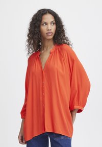 Blusa arancione fluttuante con scollatura a V, chiusura con bottoni davanti e maniche a tre quarti. Realizzata in tessuto leggero con una superficie strutturata.
