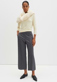 Lichtcrème lange mouwen top, grijze wijde broek en een chunky crèmekleurige sjaal. De outfit heeft een combinatie van zachte texturen en op maat gemaakte lijnen. Zwarte instap schoenen.