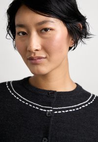 Sötét szürke kötött kardigán, kerek nyakkal, fehér cérnavonalakkal és fekete gombokkal az elején. Puha tapintású.