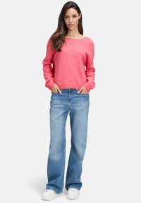 Eine Frau steht mit den Händen in den Taschen, trägt einen pinken Pullover, helle, weit geschnittene Jeans und weiße Sneakers vor einem schlichten weißen Hintergrund.