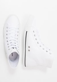 Weiße Leinwand-Hochschnitt-Sneaker mit flachen Schnürsenkeln, silbernen Ösen und schwarzer Gummisohlen-Akzent. Glatte Textur mit Logo auf dem Innenfutter.