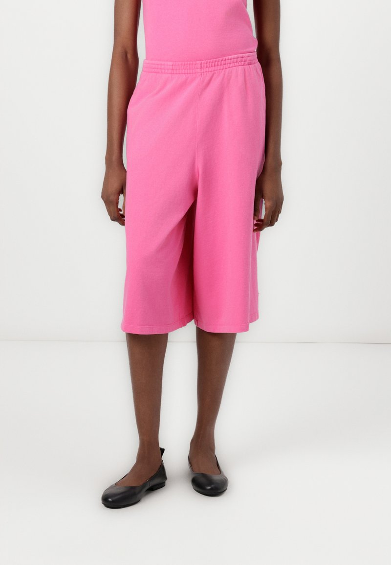 Ροδοκόκκινες βαμβακερές βερμούδες με ελαστική μέση, που φτάνουν μέχρι το μεσαίο γόνατο. Λείο υφή, άνετη εφαρμογή, συνδυασμένες με μαύρες επίπεδες παπουτσάκια.