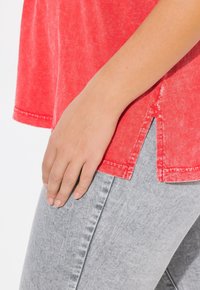 Rode katoenen T-shirt met een relaxte pasvorm, voorzien van een zijsplit, gedragen over lichtgrijze denim jeans met een vervaagde textuur en subtiele stikdetails.