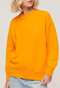 Sudadera de color naranja brillante con cuello redondo y puños de canalé, hecha de una tela suave. Presenta un pequeño logotipo bordado en el dobladillo.