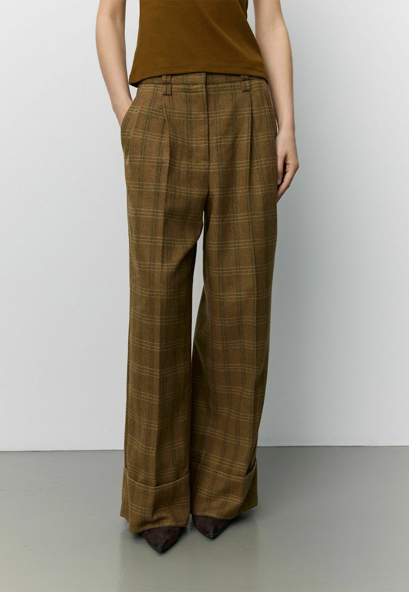 Personne portant un pantalon large à carreaux marron avec des ourlets retroussés et un haut marron sans manches, debout sur un sol gris contre un mur blanc.