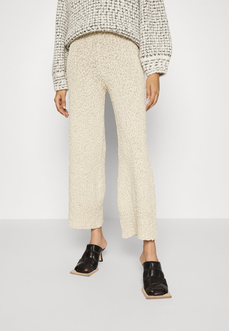 Cremfarbene, gestrickte weit geschnittene Hose mit strukturiertem Finish, kombiniert mit offenen schwarzen Absatzmules. Der Pullover hat ein kariertes Muster in Grautönen.