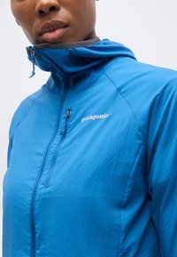 Άνδρας που φοράει μπλε ζακέτα Patagonia με φερμουάρ και γιακά που είναι κλειστός μέχρι τη μέση, κοιτά μπροστά με απλό φόντο.