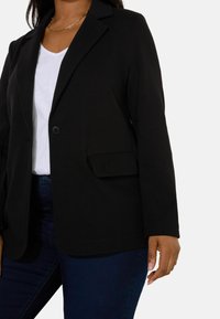 Black blazer featuring a textured fabric, single button closure, and front pocket, layered over a white t-shirt, paired with dark jeans.