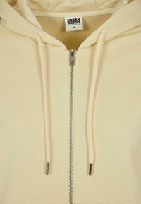Sudadera con cremallera de color amarillo claro, fabricada con material orgánico. Cuenta con capucha con cordones, cremallera de metal y puños acanalados. La etiqueta dice "URBAN CLASSICS."