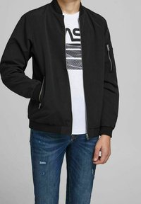 Svart bomberjacka i slät tyg, med dragkedjefickor och ribbade ärmslut. Bärs över en vit t-shirt med grafisk design och blå jeans.