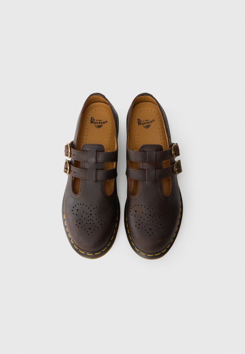 Un paio di scarpe T-bar in pelle marrone scuro di Dr. Martens, con doppie cinghie a fibbia e un design a cuori perforato sulla punta, viste dall'alto.