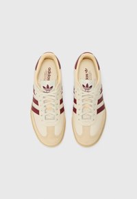 Cremefarvede sneakers med maroon striber, suede detaljer og snørebåndsdesign. Sålen er lys beige med teksturerede mønsterdetaljer.