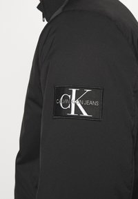 Giubbotto impermeabile nero con una grande toppa rettangolare con il testo "CALVIN KLEIN JEANS" e un logo "CK" in bianco. Texture liscia.