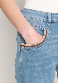 Světlé modré džínové kalhoty s boční kapsou, vybavenou kontrastním krémovo-hnědým zipem. Tkanina má měkkou, mírně opotřebovanou strukturu.