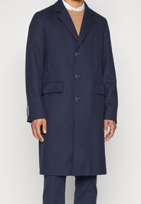 Mörkblå ullrock med spetsig krage, två framfickor, sex knappar och en skräddarsydd passform. Slät textur med en klassisk design.