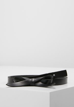 Ceinture en cuir noire avec un détail nœud, enroulée sur une surface blanche sur un fond gris clair.