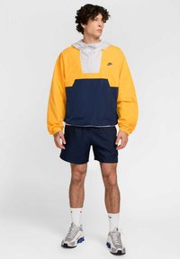 Chaqueta de pullover Nike en amarillo y marino, con capucha gris, bolsillo frontal y puños elásticos. Combinada con pantalones cortos marinos y zapatillas deportivas grises.