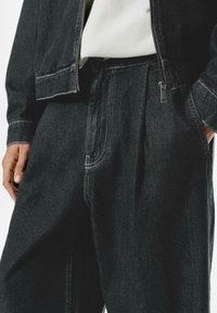 Black denim jacket and trousers, featuring light stitching and a zip detail. The fabric has a textured finish with a relaxed fit design.
