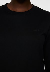 Svart sweatshirt med ribbad krage och en matt svart logga på bröstet, tillverkad av ett mjukt, texturerat material. Enkel design.