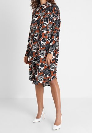 Frau trägt ein braunes, langärmliges Kleid mit Blumenmuster und weiße, spitz zulaufende High Heels, steht vor einem schlichten Hintergrund.