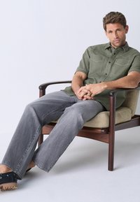 Camisa de manga corta verde con botones y dos bolsillos en el pecho, pantalones de mezclilla gris y sandalias negras, sentado en una silla de madera con un cojín beige.