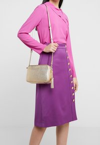 Bolso cruzado metálico dorado con acabado texturizado, correa de cadena y adorno de borlas, combinado con una blusa rosa de manga larga y una falda morada con botones.
