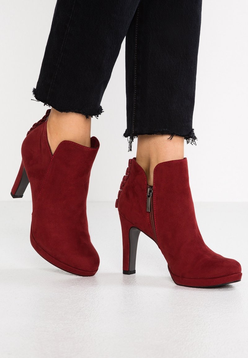 Bottes rouges en suédé à talons montant jusqu'à la cheville avec fermetures éclair latérales, portées avec un pantalon noir court à ourlet effiloché sur un fond de sol blanc.