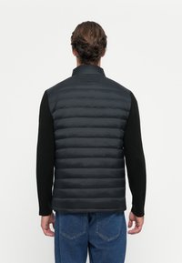 Black puffer vest with horizontal quilted sections. High collar and dark knitted sleeves. Worn over blue denim jeans.