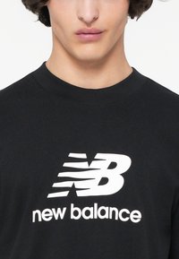 Juodos medvilnės marškinėliai su dideliu baltu "NB" logotipu ir "new balance" tekstu drąsiu, moderniu šriftu. Paprasta apvalios kaklo dizaino.