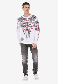 Witte lange mouwen t-shirt met zwart en rode grafische print, met sterren en ingewikkelde ontwerpen, gecombineerd met grijze jeans en witte sneakers.