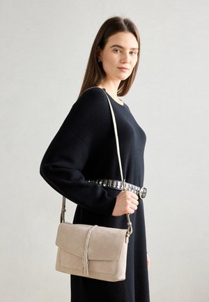 Vrouw met een zwarte jurk en een gespikt riem houdt een beige suède schoudertas met gevlochten kwastdetail vast.