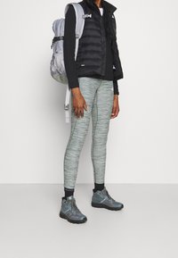 Schwarze Puffer-Weste über einem Langarmoberteil, kombiniert mit hellgrünen, strukturierten Leggings und grauen Wanderschuhen. Ein grauer Rucksack rundet das Outfit ab.